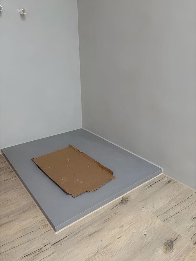 Ein minimalistischer Raum mit grauen Wänden und einem glatten grauen Podest, auf dem ein Stück Pappe auf einem Holzboden liegt.