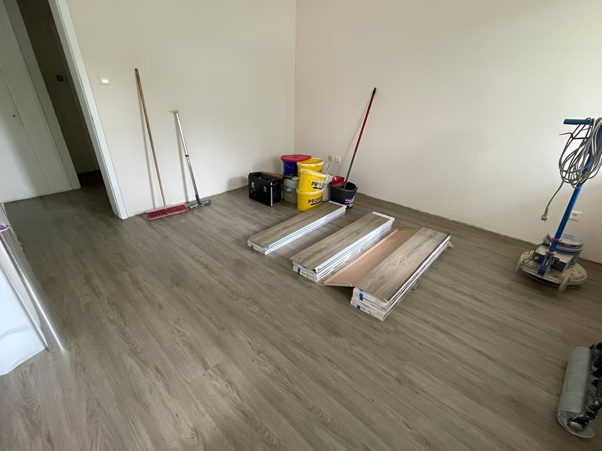 Ein leerer Raum mit grauem Laminatboden, Reinigungswerkzeugen, Eimern und Verlegewerkstoffen, die für die Renovierung bereitstehen.